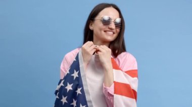 Gülümseyen mutlu genç kadın Amerikan bayrağı taşıyan günlük kıyafetler giyiyor ve mavi arka planda izole edilmiş kameraya bakıyor.