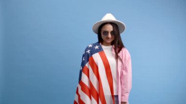 Pembe gömlekli genç esmer kadın stüdyoda mavi arka planda Amerikan bayrağı ve 100 dolar nakitle poz veriyor. Paranın kokusu.