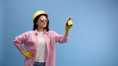 Sarı kasklı ve eldivenli bir kadın mavi stüdyo arka planına işaret ediyor ve işaret ediyor.