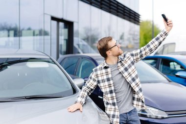 Mutlu adam yeni araba aldıktan sonra akıllı telefondan selfie çekiyor. Telefondaki video bağlantısıyla satın alınan arabayı gösteren adam..