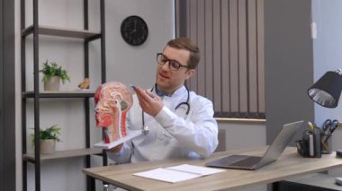 A young attractive otolaryngologist doctor shows a model of the human head and tells the patient about the structure of the respiratory system