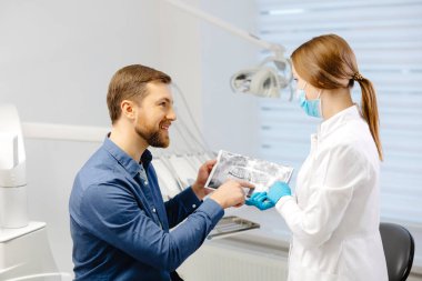 Young attractive man visiting dentist, sitting in dental chair at modern light clinic. Young woman dentist holding x ray image