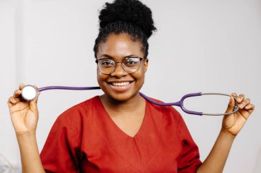 Kırmızı önlüklü, gözlüklü, gülümseyen bir kadın doktor stetoskop tutuyor..