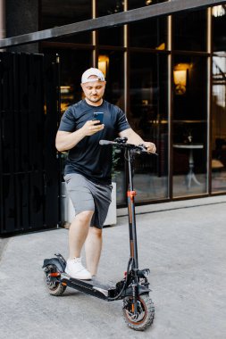 Modern binanın yanındaki caddede elektrikli scooterlı akıllı telefon kullanan genç adam.