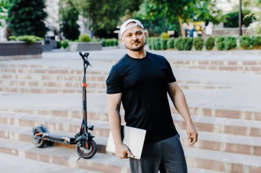 Elinde dizüstü bilgisayarı olan yakışıklı bir adam elektrikli scooter 'ın yanında duruyordu. Aktif yaşam tarzı, sırt çantalı gezgin. Ekoloji ve temiz ekolojik ulaşım biçimi
