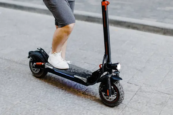 Şortlu ve spor ayakkabılı bir adamın şehir caddesinde dikilirken elektrikli scooter 'a bindiği görüntüler. Ekoloji ve temiz ekolojik ulaşım biçimi