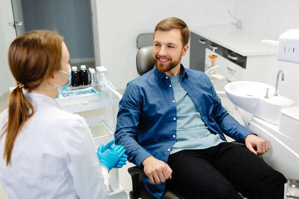 Blonde female dentist in dental office talking with male patient and preparing for treatment. Handsome bearded man in dentist chair looking at his doctor with smile.