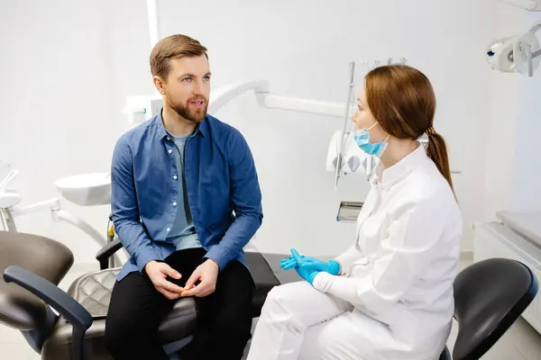 Blonde female dentist in dental office talking with male patient and preparing for treatment. Handsome bearded man in dentist chair looking at his doctor with smile.