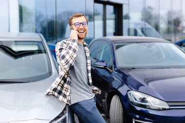 Yakışıklı genç adam cep telefonuyla konuşuyor ve arabasına yaslanırken gülümsüyor..