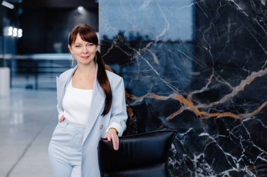 Açık mavi takım elbiseli bir kadın siyah mermer bir duvarın önünde duruyor. O gülümsüyor ve siyah bir deri ofis sandalyesinin arkasına yaslanıyor. Uzun kahverengi saçları at kuyruğu şeklinde geri çekiliyor..