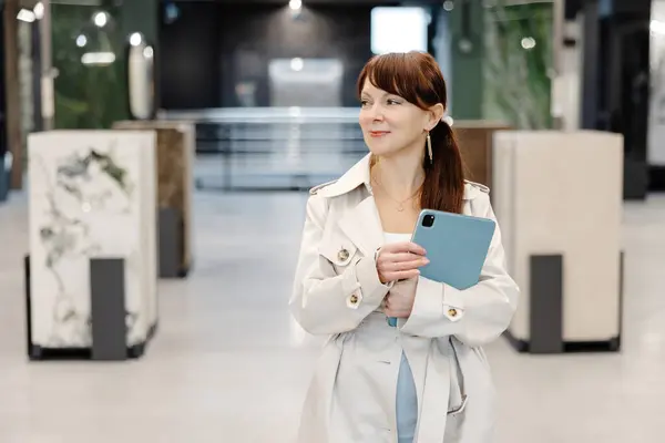 stock image A woman in a white coat is holding a blue tablet while standing in a showroom. The showroom has large, white tiled walls and a modern, minimalist design.