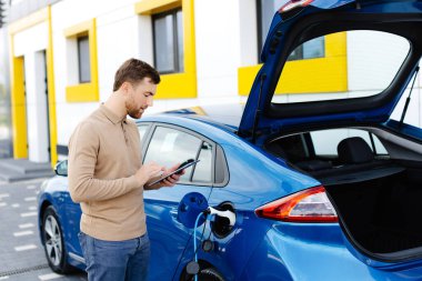 Genç bir adam bir istasyonda durup tablet tutarken elektrikli bir arabayı şarj ediyor..