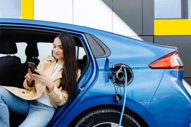 İş kadını içeride oturmuş elektrikli arabayı şarj ediyor ve modern akıllı telefonu kullanıyor. Teknoloji ve ulaşım kavramı