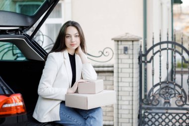 Kız arabanın bagajında oturuyor, elinde kutular, gülümsüyor, kameraya bakıyor.