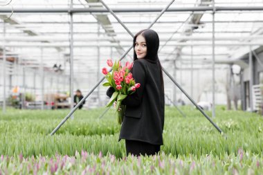 Serada duran ve elinde çiçek açan laleler tutan genç bir kadın..