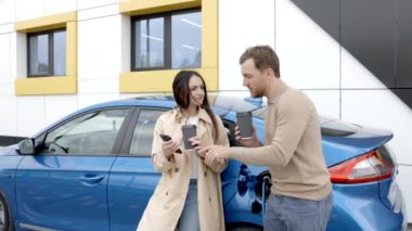 Elektrikli araba ile seyahat eden genç bir çift şarj istasyonunda durdular. Erkek arkadaşın kabloyu şarj ediyor. Sevgilisiyle konuşan, elinde fincan tutan sıcak kahve içen, gülümseyen bir adam.