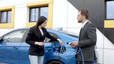 Satış müdürü genç bir müşteriye elektrikli arabanın nasıl şarj edileceğini gösteriyor. Dışarıdaki araba galerisindeki araba prizine kablo bağlıyor.