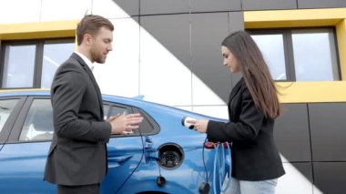 Satış müdürü genç bir müşteriye elektrikli arabanın nasıl şarj edileceğini gösteriyor. Dışarıdaki araba galerisindeki araba prizine kablo bağlıyor.