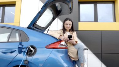 Genç bir kadın, küçük bir kamu istasyonundan şarj edilen otomobil aküsünü doldurmak için bekliyor ve otomobili şarj ederken akıllı telefon kullanıyor