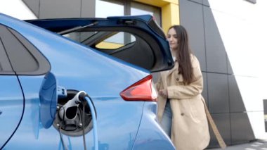 İstasyonda arabayı şarj ederken genç bir kadın elektrikli bir arabanın yanında duruyor ve bagajını bagaja koyuyor.