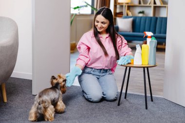 Evde temizlik malzemeleriyle masanın yanında oturan genç bir kadın ve küçük köpeği onun yanında koşuyor..