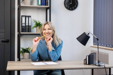 Takım elbiseli genç bir kadın ofiste masada oturuyor, elinde bir kalem tutuyor ve bir şey düşünüyor..