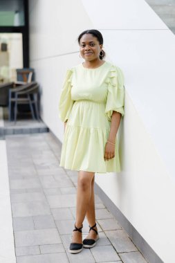 Koyu tenli bayan dışarıda gülüyor. Sarı elbiseli esmer kadın gülümsüyor ve dışarı çıkıyor..