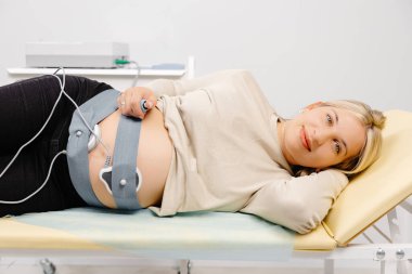 Woman pregnant checking fetal heart beat by fetal monitoring. Expectant mother with midwife adjusting sensors off cardiotocography(CTG). clipart