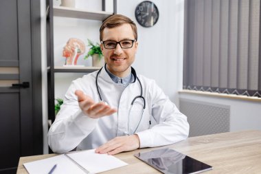 Portrait of young doctor wearing glasses and white uniform with stethoscope, speaking and consulting patient online, looking at camera, making video call, sitting at table in office.