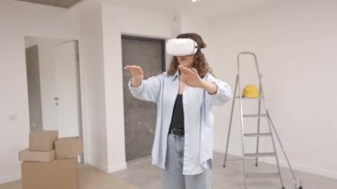 VR gözlüklü genç bir kadın modern teknolojiyi kullanarak oda tasarımı yapıyor, ellerini havada hareket ettiriyor, yenileme sırasında yeni bir dairede duruyor. Sanal gerçeklik ve onarım kavramı