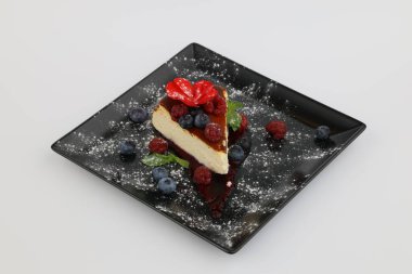 Böğürtlenli ve soslu leziz cheesecake, beyaz arka planda siyah bir tabakta..