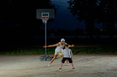 Bir baba, parlak bir ışıkla aydınlatılmış bir parkta genç oğluyla basketbol oynar.