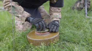 Özel takım elbiseli bir adam dedektörle çalışıyor ve patlayıcı bir cihaz buldu. Adamın biri tanksavar mayınını etkisiz hale getirmeye çalışıyor.