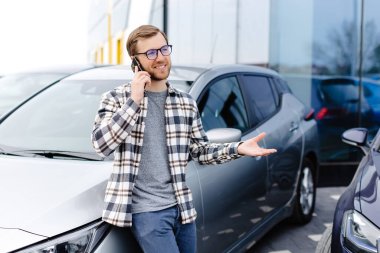 Yakışıklı genç adam cep telefonuyla konuşuyor ve arabasına yaslanırken gülümsüyor..