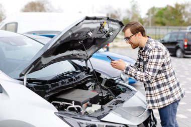 Genç adam arabanın kaputunun altını kontrol ediyor. Sorunun ne olduğunu anlamak için.