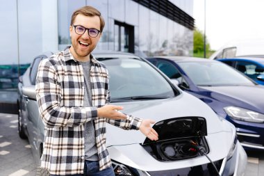 Genç bir adam yeni elektrikli arabasının yanında duruyor ve araba şarj olurken dinleniyor..