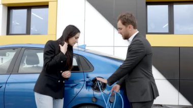 Satış müdürü genç bir müşteriye elektrikli arabanın nasıl şarj edileceğini gösteriyor. Dışarıdaki araba galerisindeki araba prizine kablo bağlıyor.