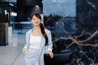 Açık mavi takım elbiseli bir kadın siyah mermer bir duvarın önünde duruyor. O gülümsüyor ve siyah bir deri ofis sandalyesinin arkasına yaslanıyor. Uzun kahverengi saçları at kuyruğu şeklinde geri çekiliyor..