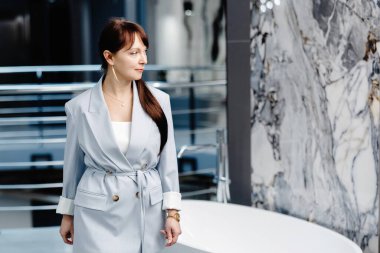 Açık gri takım elbiseli bir kadın modern bir galeride duruyor, çerçevenin sağ tarafına bakıyor. Ön planda büyük, beyaz, serbest bir küvet var. Kadının sağ eli cebinde, ve sol eli de cebinde duruyor.