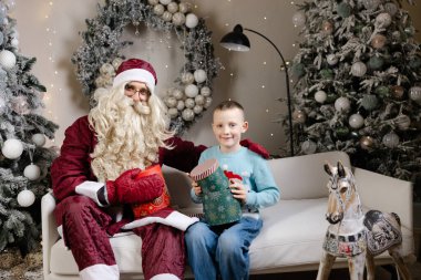 Noel Baba, dekore edilmiş bir odada beyaz bir kanepede oturan mutlu bir çocuğa Noel hediyesi veriyor.
