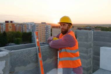 Gün batımında beton bloğa yaslanmış çelik yelek ve şapka giyen kendine güvenen inşaat işçisinin portresi.