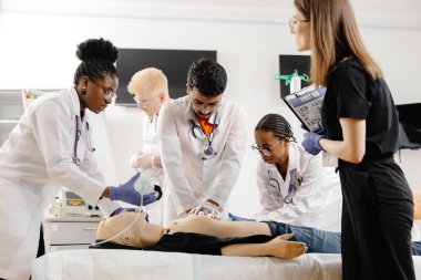 Bir grup beyaz önlüklü doktor klinik odasındaki muayene masasında oturan bir hastayla sohbet ediyorlar..