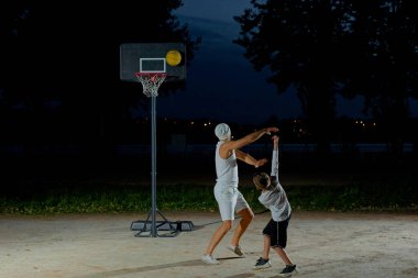 Bir baba, parlak bir ışıkla aydınlatılmış bir parkta genç oğluyla basketbol oynar.