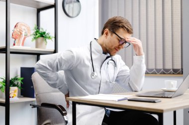 A young male doctor sits at his workplace and feels pain in his back. A doctor takes a break from his work day because of pain.