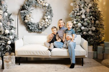 Anne ve iki oğlu Noel süslemeli bir oturma odasında beyaz bir koltukta oturuyorlar.