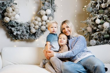 Anne ve iki oğlu Noel süslemeli bir oturma odasında beyaz bir koltukta oturuyorlar.