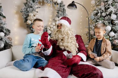 Noel Baba beyaz koltukta oturan iki süpriz çocuk ve onlara Noel ağacının yanında para veriyor.