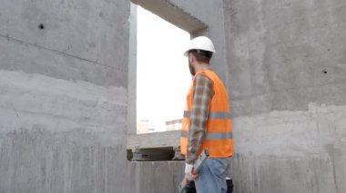 Tamirci, koruyucu miğfer ve yelek giyen ustabaşı inşaat alanında duruyor ve elinde cetvel ve alet çantası tutuyor.