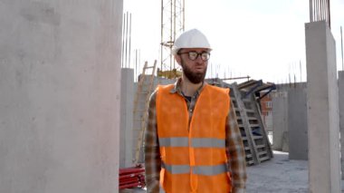 İnşaat işçisi, miğfer ve yelek giyen ustabaşı inşaat halindeki bir binada duruyor ve el hareketi gösteriyor.