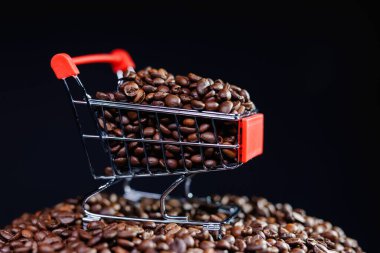 Kavrulmuş kahve çekirdekleri ile dolup taşan minyatür alışveriş arabası koyu arkaplana karşı daha fazla kahve çekirdeği yığını üzerine yerleştirilir.